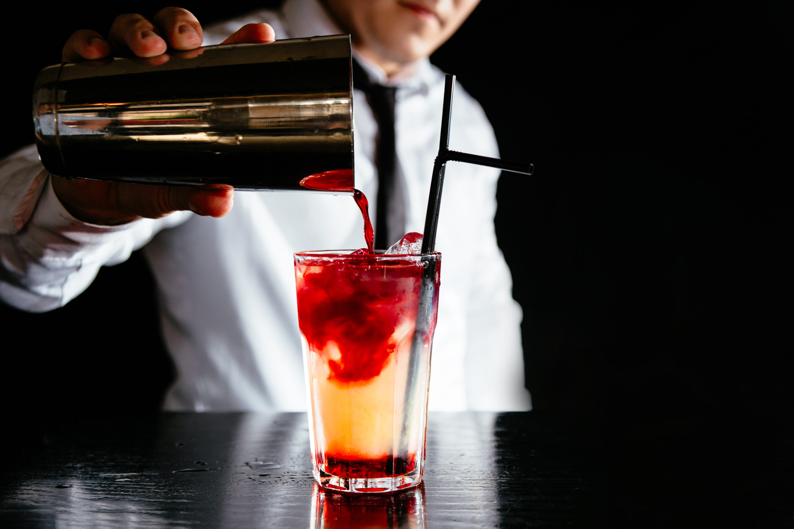 Barman makes a two colored cocktail on a black counter, black ba